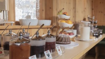 Ein Buffet mit verschiedenen Marmeladen in Glasbehältern und einem Obstteller. Im Hintergrund sind zusätzliche Snacks und Getränke sichtbar.