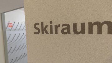 Ein Schild mit der Aufschrift "Skiraum" an einer Wand. Im Hintergrund sind Garderobenhaken sichtbar.