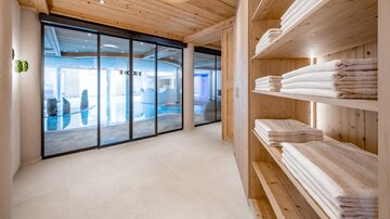 A modern wellness area with wooden-paneled walls and a clear view of a pool. A shelf with folded towels provides a relaxing atmosphere.