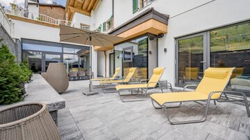 A modern terrace with yellow loungers and a sun umbrella. In the background, large windows and an elegant residential building can be seen.
