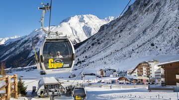 A cable car travels through a snow-covered landscape with high mountains. In the foreground, buildings and ski slopes are visible.