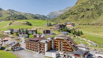 A picturesque mountain landscape with several buildings and green meadows. The sun is shining and the sky is clear.