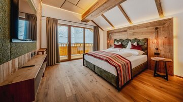 A modern bedroom with wood decor and large windows. The cozy bed is decorated with red and green pillows and offers a view of the mountains.