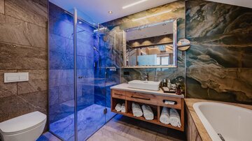 A modern bathroom with a glass shower and an elegant bathtub. The wooden cabinet with towels and the colorful stone on the wall add a stylish look.