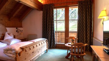A cozy bedroom with wooden details and two windows. There is a bed, a table with chairs, and plenty of daylight.